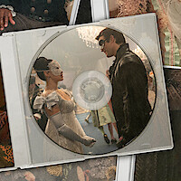A collage of historical costumes and accessories surrounds a CD case, which shows a masked ballroom scene with two people dressed in elegant, vintage attire facing each other.