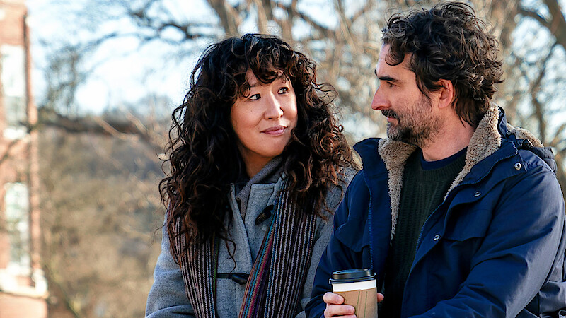 Two people standing outdoors in winter coats, looking at each other and smiling. One is holding a coffee cup. Background includes bare trees and a building, suggesting a chilly day in a park or campus setting.