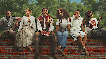 Six young adults sit on a red brick wall outdoors at Hillman College, dressed in casual and vintage clothing, smiling and relaxed, with green trees in the background.