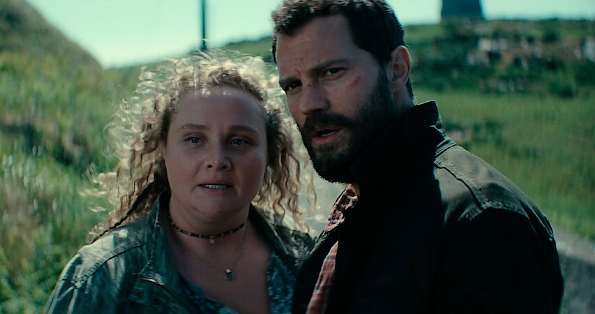 Jamie Dornan as Elliot and Danielle Macdonald as Helen stand together on a grassy hill in season 2 of 'The Tourist'