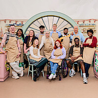Group of twelve people in aprons posing together inside a colorful baking tent, likely contestants for a baking competition, with kitchen equipment and pastel decor in the background.