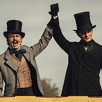 Two men in vintage attire and top hats stand on a platform outdoors, holding hands victoriously, conveying a celebratory and triumphant mood under a cloudy sky, likely at a historical event or reenactment.