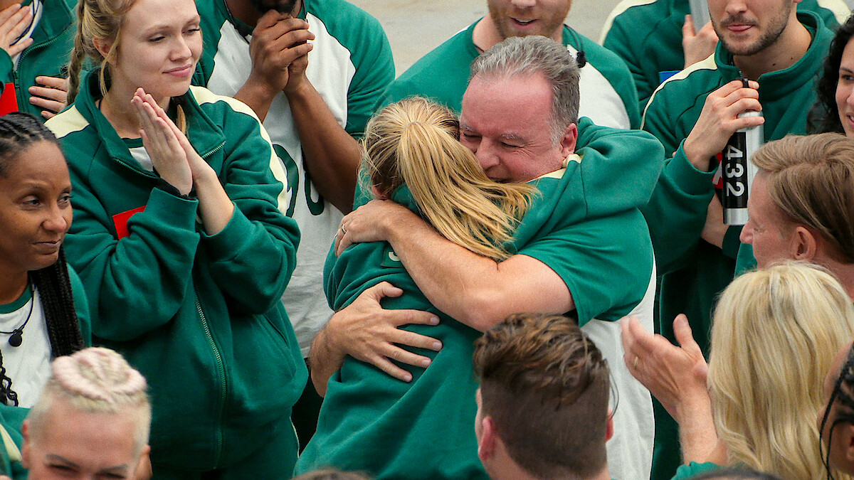 A group of people in green tracksuits celebrate as two participants share a joyful, emotional hug in the center. Others around them clap and smile, creating a supportive and uplifting atmosphere.