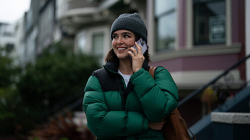 Woman in a green puffer jacket and gray beanie smiling while talking on a phone outside near colorful residential buildings on a cloudy day.