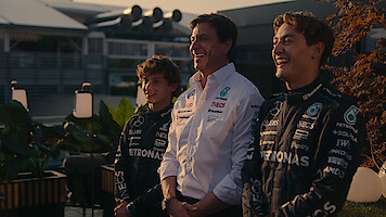 Three people, two in black Mercedes Formula 1 racing suits and one in a white team shirt, stand smiling outdoors at a race venue with plants and a modern building in the background during daylight.