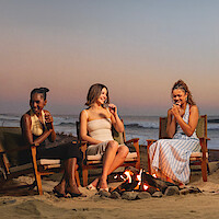 Three women sit around a fire pit on a sandy beach at sunset, laughing and enjoying drinks, with waves in the background and wooden chairs arranged in a circle.