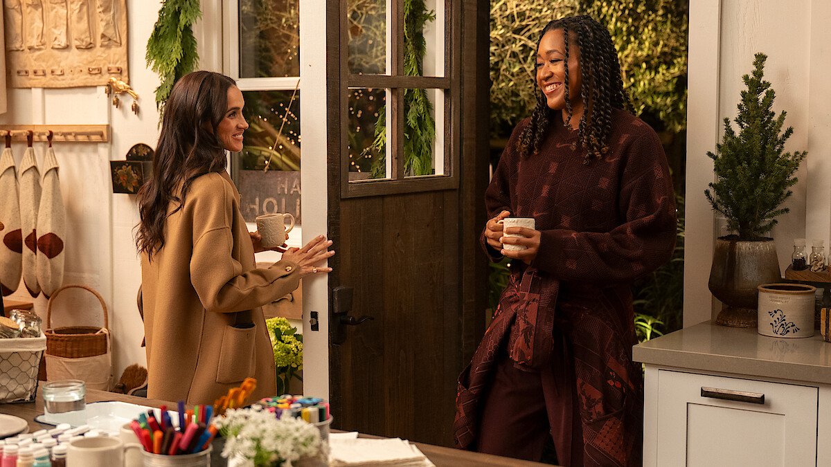 Two women smiling and talking in a cozy kitchen decorated for the holidays, holding mugs. Warm lighting, craft supplies on the counter, and greenery create a welcoming, festive atmosphere.