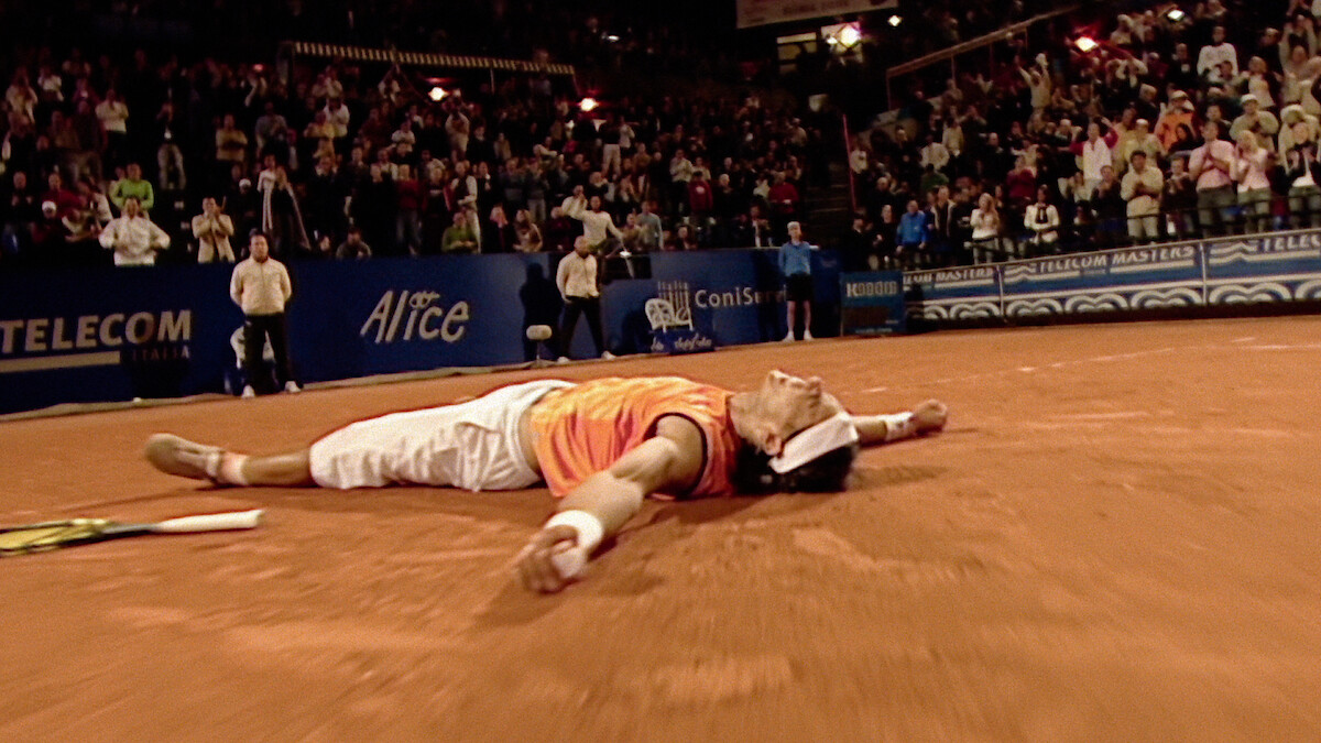 Tennis player lying on back on clay court, arms spread, appearing exhausted or celebrating victory, with tennis racket beside and cheering crowd in evening stadium.
