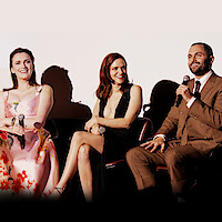 From left to right, Charlotte Ritchie, Madeline Brewer, and Penn Badgley sitting on a panel, each holding a microphone and smiling. They are dressed in formal attire.