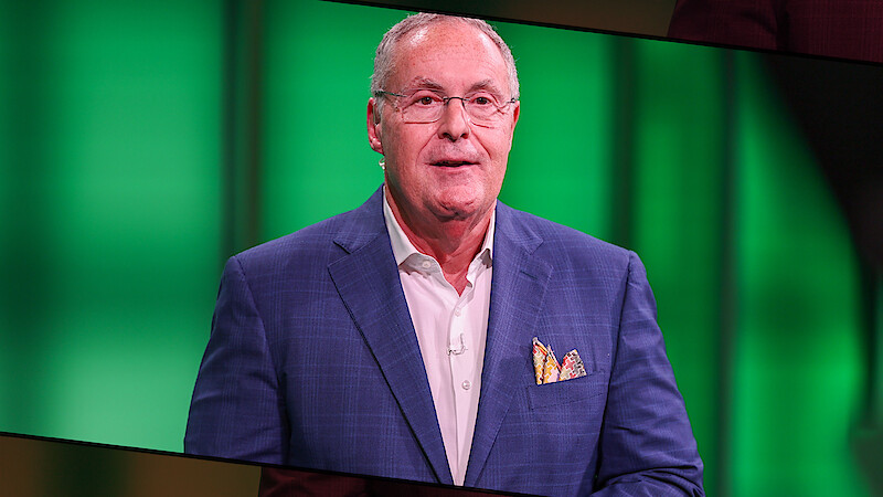 An older man in a blue blazer with a pocket square stands in a brightly lit studio with a green background.