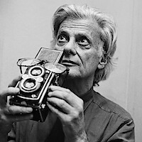 Older man with gray hair in a collared shirt holding a vintage twin-lens reflex camera, gazing thoughtfully upward in an indoor setting with neutral background. Black and white photo, studio light ambiance.