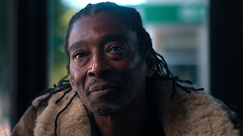 Man with dreadlocks sitting indoors, wearing a shearling jacket, soft natural light on his face, background blurred with green and white tones, calm and contemplative mood.