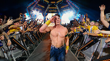 Shirtless wrestler saluting and walking down a ramp, surrounded by cheering fans at a brightly lit arena event, with colorful lights and stage setup in the background.