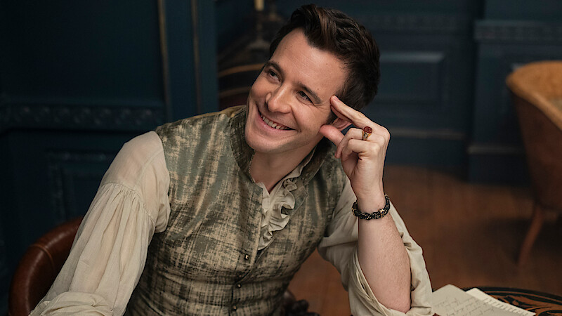 Man in period clothing sitting at a wooden desk, smiling and looking to the side in a warmly lit, elegant room with dark paneling and papers on the desk.