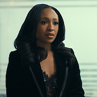 Woman with long, styled hair wearing a black blazer and lace top, sitting indoors in a modern, minimalistic environment with a neutral background, looking attentive and composed.