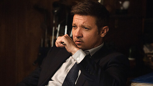 A man in a suit sits indoors, appearing deep in thought with his hand near his mouth, in a dimly lit room with wooden walls and blurred objects in the background.