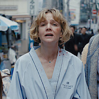 Carey Mulligan as Lindsay Crane-Martin with short blonde hair, wearing a blue robe with red stains, appears distressed while walking down a busy city street surrounded by people and urban storefronts.