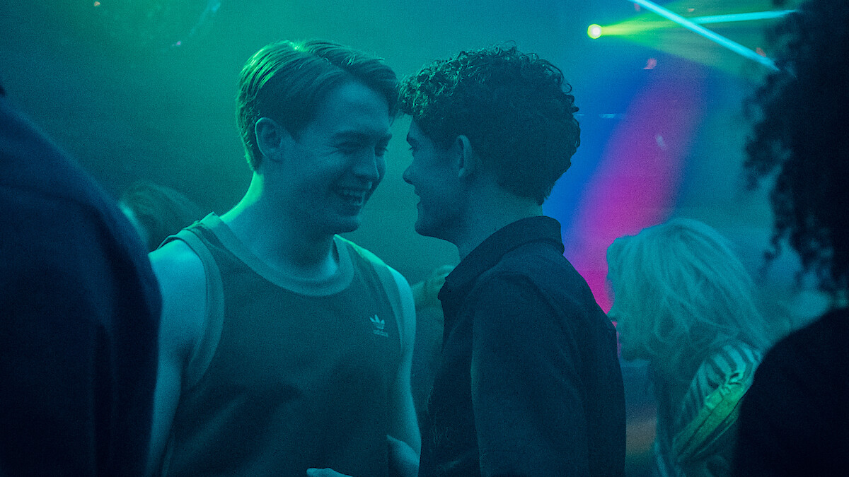 Two teenagers in a cozy, dimly lit bedroom, one sitting on the floor using a laptop, the other sitting on a bed watching. The room has string lights, posters, and personal items, creating a relaxed, lived-in atmosphere.