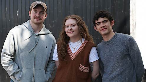 Three young adults standing side by side outdoors in front of a dark wooden wall, smiling at the camera. Casual clothing, natural lighting, and a relaxed atmosphere.
