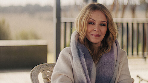 Woman with long blond hair wearing a cozy cream sweater and a thick purple-gray scarf, sitting outdoors with blurred fence and trees in the background on a soft, overcast day.