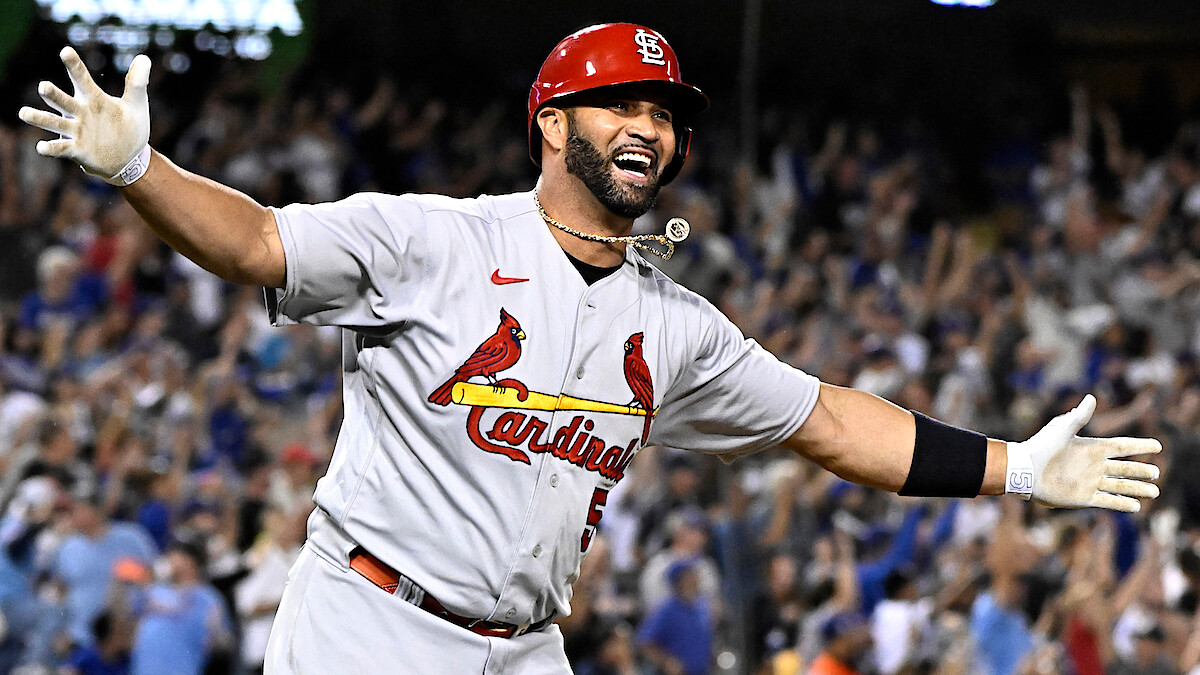 Albert Pujols in a St. Louis Cardinals uniform celebrating on field with arms outstretched, smiling, with cheering crowd in stadium background during night game.