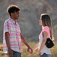 Two people, a young man and woman, stand outdoors in a sunny, rocky landscape, holding hands and looking at each other, both wearing casual clothes with natural light highlighting their faces.