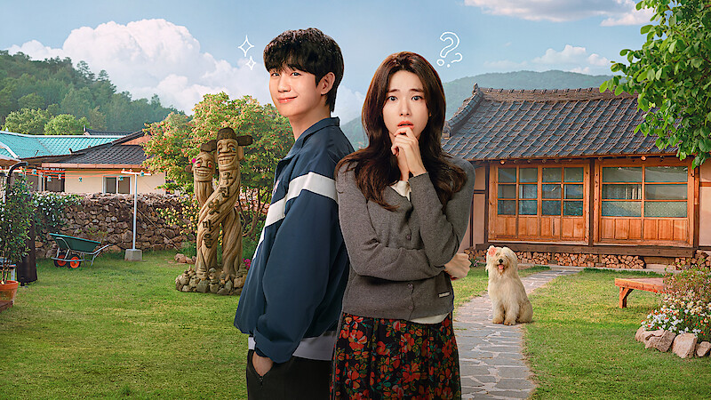 A young man and woman stand in a sunny, rural yard with traditional wooden house, lawn, garden statues, and a fluffy dog, mountains in the background, blue sky with clouds, both subjects displaying thoughtful and playful expressions.