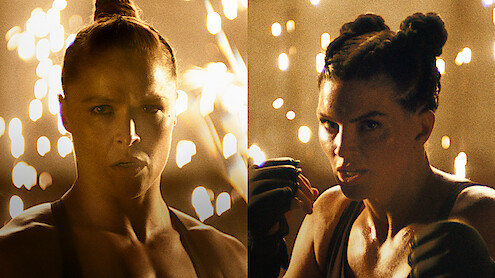 Ronda Rousey and Gina Carano with intense expressions face forward in a dramatic, dimly lit environment with sparks flying in the background, both wearing athletic gear and preparing to fight.