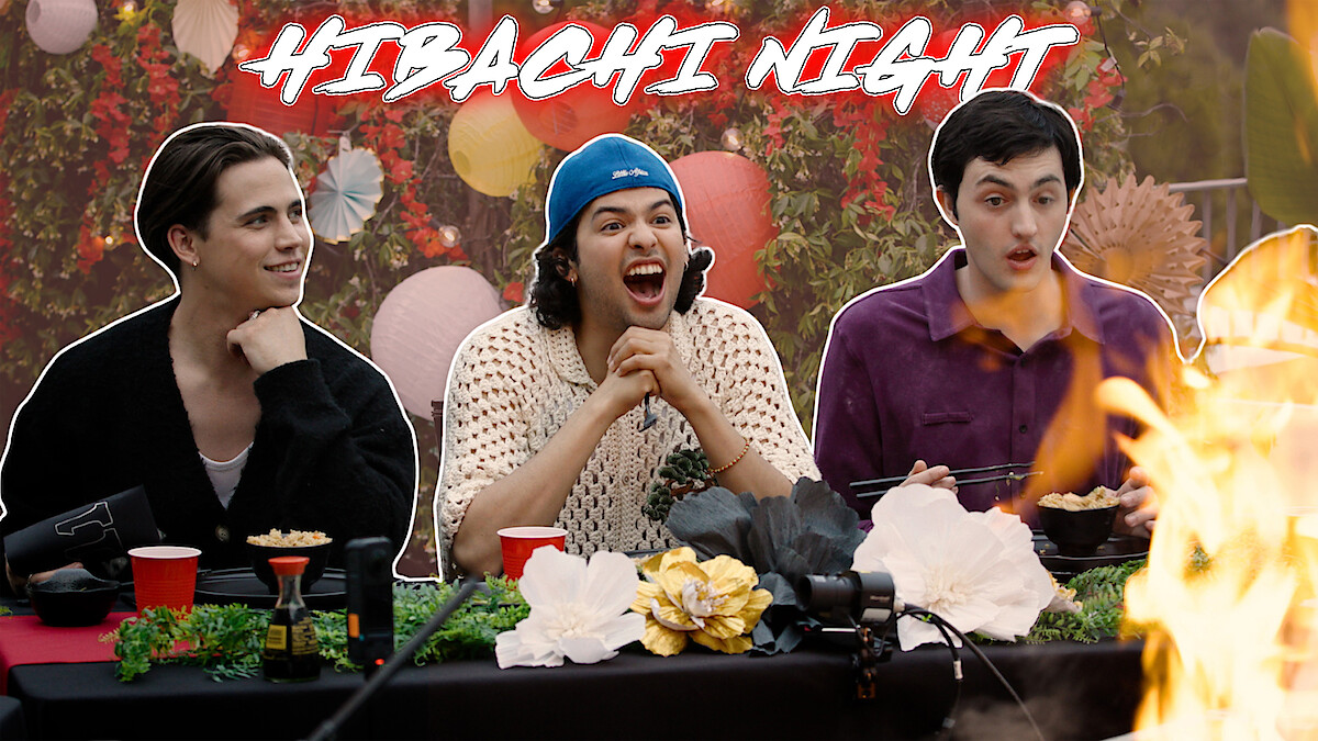 From left to right, Tanner Buchanan, Xolo Maridueña, and Gianni DeCenzo are seated at a dinner table, with flames rising dramatically on the right side.