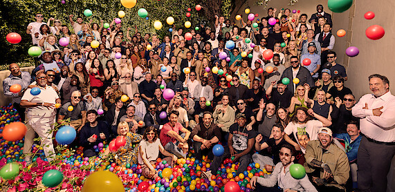 Large group of people of various ages gathered outdoors, posing in a ball pit with colorful plastic balls bouncing in the air; casual and joyful setting with trees and a wall in the background.