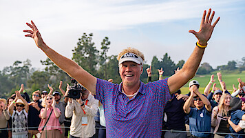 Smiling golfer with raised arms celebrates on a golf course, surrounded by cheering spectators and trees in the background, under a partly cloudy sky.