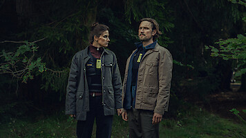 Two people wearing badges and jackets stand side by side in a dense, green forest, holding hands and looking in opposite directions, suggesting a serious or investigative scenario in a natural outdoor setting.