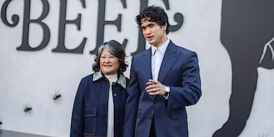 Two people stand together, smiling, in front of a wall with large black text and silhouettes as part of an event backdrop. The man wears a dark suit and white shirt, while the woman wears a dark jacket with a light collar.
