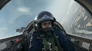 Pilot in cockpit flying a jet fighter, wearing helmet and oxygen mask, with intense expression; another jet visible outside, sky and landscape blurred from high speed and aerial maneuver.