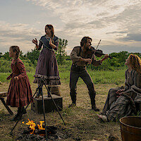 A man plays violin while three women and girls in period clothing laugh and dance around a campfire in a grassy outdoor rural setting at sunset, evoking a joyful, historical atmosphere.