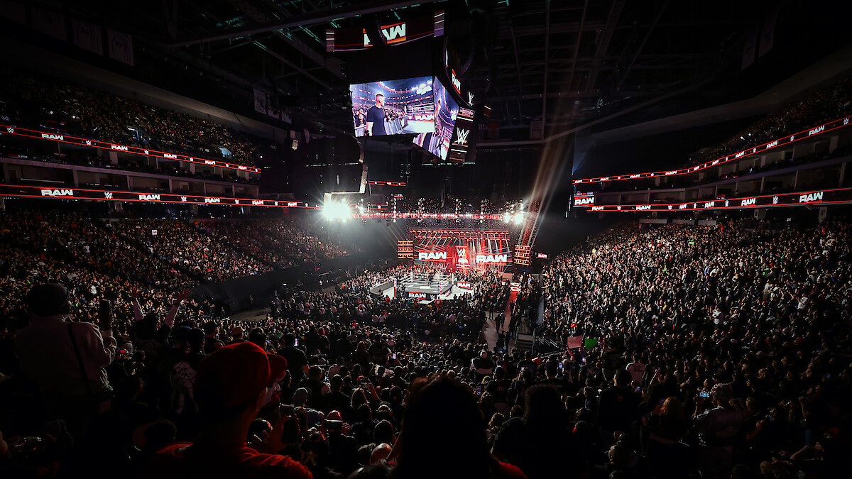 Large, packed arena hosting a WWE wrestling event with bright lights, digital screens, and enthusiastic crowd surrounding a central ring under the arena’s high ceiling.
