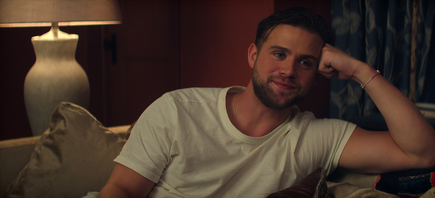 Leo Woodall as Vladimir in a white t-shirt relaxing on a sofa in a cozy, dimly lit living room with pillows, a lamp, and curtains in the background.