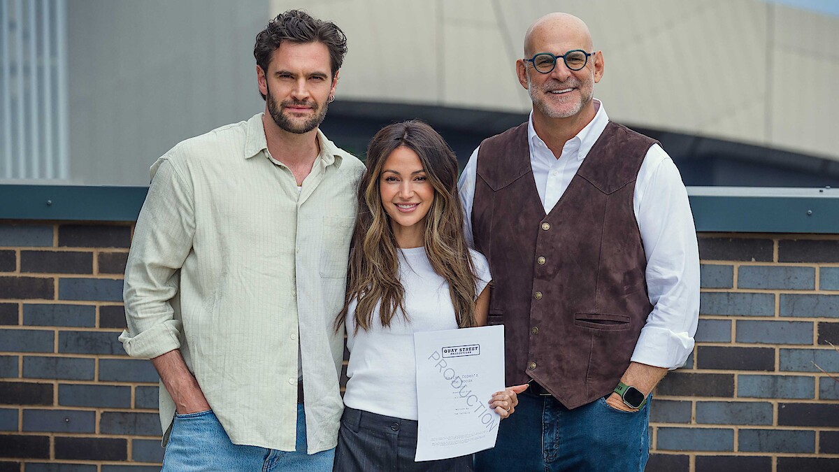 Three people standing together outdoors by a brick wall, one person in the middle holding a document and smiling, all appear happy in a casual urban setting with a large building in the background.