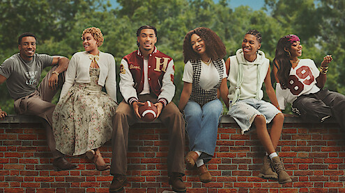 Six young adults sit on a brick wall outdoors, smiling and relaxed. Trees and blue sky in background. One holds a football; they wear casual, varied outfits, indicating a friendly, laid-back group setting.