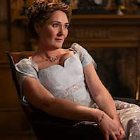 Woman in a light blue Victorian-era dress sits in an ornate wooden chair, posed thoughtfully in an elegant room with a carved wooden fireplace and classical decor in the background.