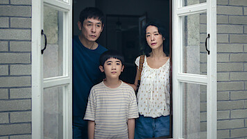 A man, woman, and young boy stand closely together inside a house, looking out through an open window with white frames. They appear serious, framed by gray brick walls and soft indoor lighting.
