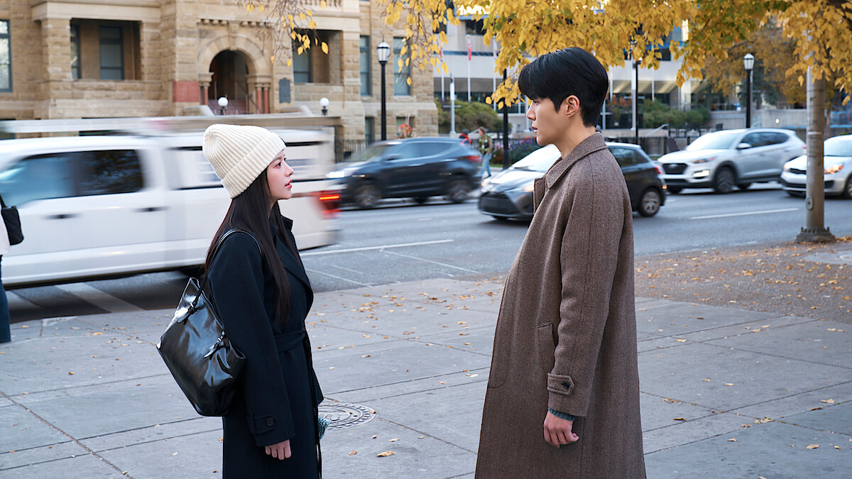Two people stand facing each other on a city sidewalk lined with autumn trees, wearing coats, with blurred cars and historic buildings in the background.