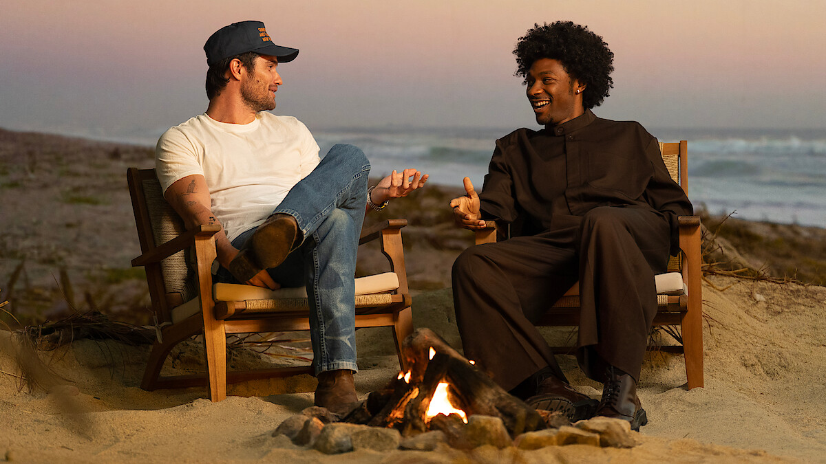 Two men sitting in chairs on a sandy beach at sunset, talking and smiling by a small campfire with the ocean and waves in the background.