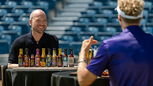 A smiling man sits at a table facing another person with several hot sauce bottle between them.