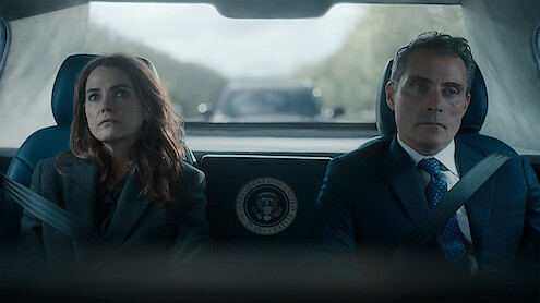 Two serious people, a woman and a man in formal attire, sit side by side in the backseat of a car with presidential seal, wearing seatbelts, with a blurred road and convoy vehicle visible through the rear window.