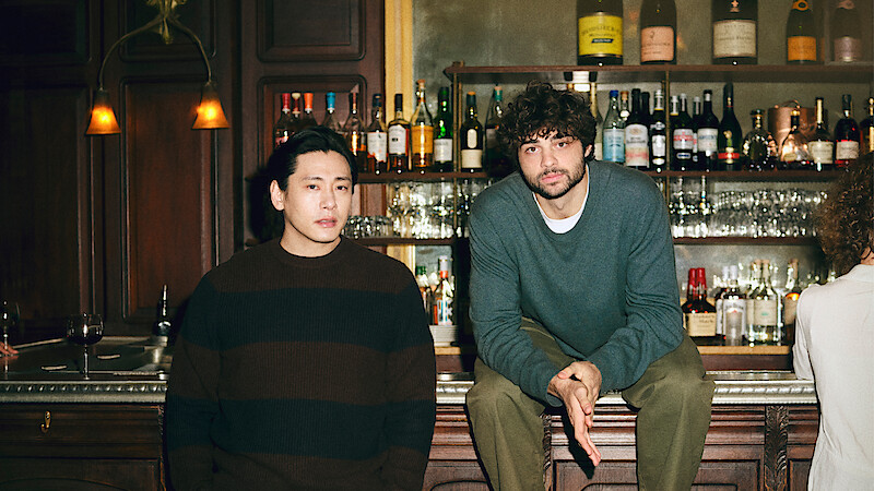 Teo Yoo leans against, and Noah Centineo sit atop the bar looking directly to camera.