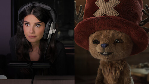 A woman with long dark hair wearing large headphones speaks into a microphone in a studio; beside her, a smiling animated creature with antlers and a large red hat stands in a warmly lit environment.