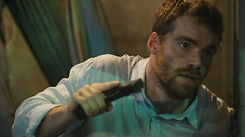 Gabriel Basso as Peter Sutherland leans on a wall holding a gun in Season 2 of 'The Night Agent'