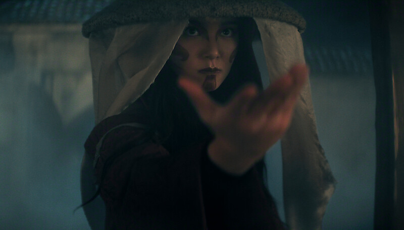 Kiawentiio as Katara stands in a misty, dimly lit setting with an outstretched hand, wearing a headdress with fabric strips, suggesting a historical or fantasy environment.
