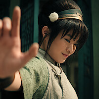 Miyako as Toph, with long hair in a bun, wearing a headband and traditional attire, smiling gently, extends hand outward confidently.